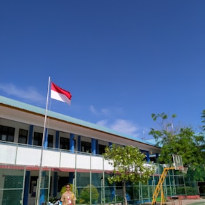 A school courtyard with a flagpole displaying a red and white flag. A person in a uniform stands near the flag. The school building in the background has a light blue roof and is surrounded by a green fence. Lush green trees and clear blue sky add to the scene.