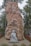 A large stone monument with engraved Vietnamese text stands prominently. In front of the stone is a small stone statue of a monk or religious figure holding a broom. The area is surrounded by greenery and red flowers, and there is a car visible in the background.