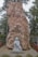 A large stone monument with engraved Vietnamese text stands prominently. In front of the stone is a small stone statue of a monk or religious figure holding a broom. The area is surrounded by greenery and red flowers, and there is a car visible in the background.