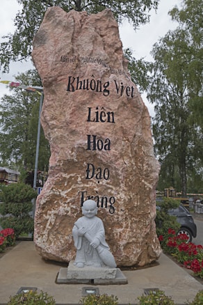 A large stone monument with engraved Vietnamese text stands prominently. In front of the stone is a small stone statue of a monk or religious figure holding a broom. The area is surrounded by greenery and red flowers, and there is a car visible in the background.