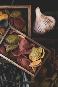 A wooden tray filled with colorful, textured pieces of dried pasta, featuring shades of red, green, yellow, and blue. The tray is set against a dark background, with a bulb of garlic placed nearby. The textures and colors create a rustic and artisanal feel, while the natural lighting enhances the setup.
