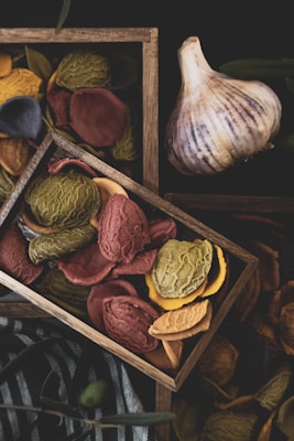 A wooden tray filled with colorful, textured pieces of dried pasta, featuring shades of red, green, yellow, and blue. The tray is set against a dark background, with a bulb of garlic placed nearby. The textures and colors create a rustic and artisanal feel, while the natural lighting enhances the setup.