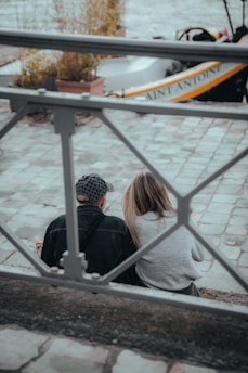Two people sit close together on a riverside pathway, facing away. The paved ground has a light, muted color. A gray metal railing runs horizontally in the foreground. In the background, a partially obscured boat with 'Saint Antoine' written on it, and potted plants can be seen.
