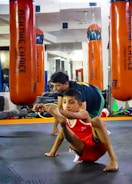 A child in athletic wear is practicing a balance exercise on the gym floor, while an instructor or coach assists nearby. Large orange punching bags are visible in the mirrored background, reflecting the gym setting.