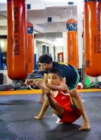 A child in athletic wear is practicing a balance exercise on the gym floor, while an instructor or coach assists nearby. Large orange punching bags are visible in the mirrored background, reflecting the gym setting.