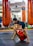 A child in athletic wear is practicing a balance exercise on the gym floor, while an instructor or coach assists nearby. Large orange punching bags are visible in the mirrored background, reflecting the gym setting.