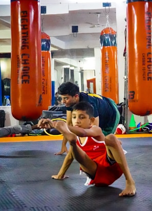 A child in athletic wear is practicing a balance exercise on the gym floor, while an instructor or coach assists nearby. Large orange punching bags are visible in the mirrored background, reflecting the gym setting.