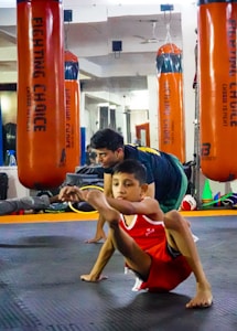 A child in athletic wear is practicing a balance exercise on the gym floor, while an instructor or coach assists nearby. Large orange punching bags are visible in the mirrored background, reflecting the gym setting.