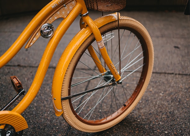 Close-up view of a vintage-style bicycle with a vibrant orange color and intricate geometric patterns on its frame. The bike features a wicker basket attached at the front and smooth tires, suitable for urban riding.