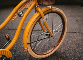Close-up view of a vintage-style bicycle with a vibrant orange color and intricate geometric patterns on its frame. The bike features a wicker basket attached at the front and smooth tires, suitable for urban riding.