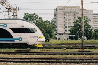 A modern train is stationed at a railway platform near a sign that reads 'Zagreb Glavni Kolodvor'. The train has a sleek white design with blue and black accents. In the background, there is a multi-story building with yellow window frames surrounded by trees and grassy areas.