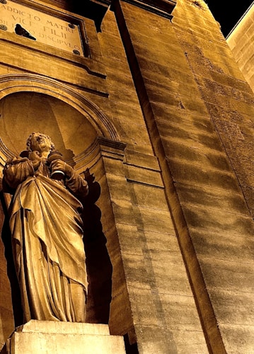 A stone statue of a robed figure holding a scroll is set within a niche in an ornate, textured wall. Above the statue is a carved inscription. The warm lighting accentuates the golden hues and the architectural details of the structure.