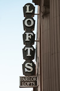 A vertical vintage-style sign attached to the side of a building displays the word 'LOFTS' in large, white capital letters on a dark background, with an additional smaller sign below reading 'PARLOR LOFTS'. The building exterior shows detailed columns and architectural elements.