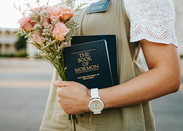 A person is holding a copy of 'The Book of Mormon' along with a bouquet of pink roses. The individual is wearing an olive green dress and a white lace-sleeved top, accessorized with a silver wristwatch.