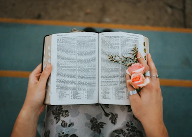 A close-up of a woman’s hands holding an open devotional book with a gold pen and blush pink flowers nearby.