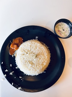A round mound of cooked white rice is served on a black plate. A small portion of orange sauce or curry is placed next to the rice. A separate small black bowl contains a yogurt-based dish with visible pieces of cucumber and onion.