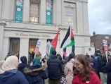 A group of people is gathered outside the St Albans Museum + Gallery. Some participants are holding flags and signs advocating for freedom in Palestine. The museum's facade is visible in the background, with large windows and banners that read 'Time for Tea.'