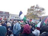 A large group of people are gathered in an urban setting, many holding Palestinian flags and signs supporting Palestine. The crowd consists of a diverse group of individuals, some wearing traditional garments. There is a sense of activism and solidarity as people engage in the demonstration, holding signs, flags, and balloons. Trees and buildings are visible in the background under a cloudy sky.