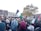 A large group of people are gathered in an urban setting, many holding Palestinian flags and signs supporting Palestine. The crowd consists of a diverse group of individuals, some wearing traditional garments. There is a sense of activism and solidarity as people engage in the demonstration, holding signs, flags, and balloons. Trees and buildings are visible in the background under a cloudy sky.