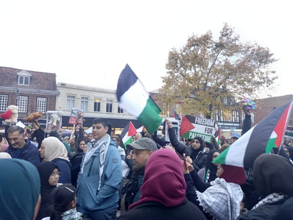 A large group of people are gathered in an urban setting, many holding Palestinian flags and signs supporting Palestine. The crowd consists of a diverse group of individuals, some wearing traditional garments. There is a sense of activism and solidarity as people engage in the demonstration, holding signs, flags, and balloons. Trees and buildings are visible in the background under a cloudy sky.