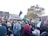 A large group of people are gathered in an urban setting, many holding Palestinian flags and signs supporting Palestine. The crowd consists of a diverse group of individuals, some wearing traditional garments. There is a sense of activism and solidarity as people engage in the demonstration, holding signs, flags, and balloons. Trees and buildings are visible in the background under a cloudy sky.
