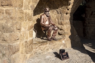A bronze statue of a seated man wearing a suit and hat is positioned against a stone wall with arches. The statue exudes the appearance of an artist or historical figure. Nearby, a small black box with a plaque indicates the name and dates associated with the figure.