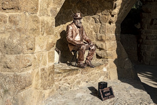 A bronze statue of a seated man wearing a suit and hat is positioned against a stone wall with arches. The statue exudes the appearance of an artist or historical figure. Nearby, a small black box with a plaque indicates the name and dates associated with the figure.