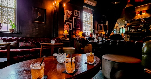 A cozy, dimly lit bar or lounge featuring leather couches, ornate lamps, and several framed pictures on the walls. On the table, there are three cocktails with straws. The atmosphere is warm and intimate, with patrons sitting and conversing in the background.