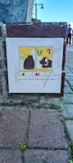 A small tile is embedded in a brick wall, featuring a simple cartoon image. The cartoon shows a black cat and a black dog looking out of adjacent windows. On the windowsill are two small birds and a potted plant. Below the illustration is a caption: 'I wish to be better acquainted with birds.' The setting includes cobblestone pavement and the lower portion of a street lamp in the background.