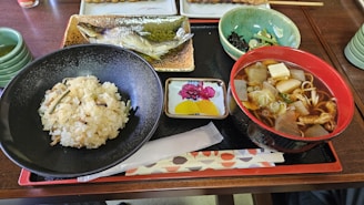 A Japanese meal served on a wooden tray includes a bowl of rice, a grilled fish presented on a rectangular plate, a bowl of soup with vegetables and tofu, and a small dish with colorful pickled vegetables. Green tea is served in a cup on the side.