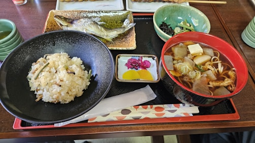 A Japanese meal served on a wooden tray includes a bowl of rice, a grilled fish presented on a rectangular plate, a bowl of soup with vegetables and tofu, and a small dish with colorful pickled vegetables. Green tea is served in a cup on the side.