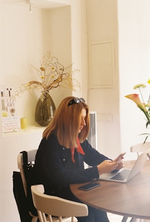 A woman with blonde hair sits at a round wooden table in a brightly lit room. She is looking at her laptop while touching her chin with her left hand. She wears a red scarf, a dark jacket, and has sunglasses resting on her head. There are various items on the table including a smartphone. In the background, on a shelf, there is a large vase with dried flowers and several pieces of paper or cards. A white wall and a window with subtle light form the backdrop, creating a cozy and warm atmosphere.