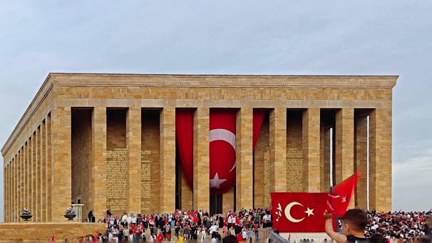 A large stone mausoleum with tall columns and Turkish flags prominently displayed. A crowd of people is gathered in front, some holding flags. The architecture is classical with inscriptions visible on the stone.