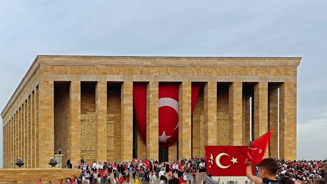 A large stone mausoleum with tall columns and Turkish flags prominently displayed. A crowd of people is gathered in front, some holding flags. The architecture is classical with inscriptions visible on the stone.