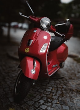 A red scooter is parked on a cobblestone path. The scooter features an Apple sticker on the front and a small emblem on the mudguard. The setting appears to be outdoors with a soft, blurred background.