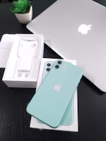 A mint green smartphone is placed on a box with the device's packaging visible, including earphones and a charger. A silver laptop with an Apple logo is also seen on a black surface next to a small green potted plant.