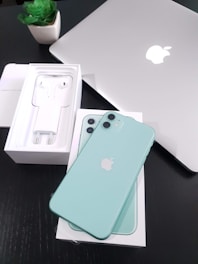 A mint green smartphone is placed on a box with the device's packaging visible, including earphones and a charger. A silver laptop with an Apple logo is also seen on a black surface next to a small green potted plant.