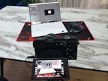 A camera positioned in front of a laptop on a marble-textured table. The camera's digital screen displays the laptop as well. A wireless mouse sits on a red and black mouse pad beside the laptop.