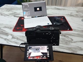 A camera positioned in front of a laptop on a marble-textured table. The camera's digital screen displays the laptop as well. A wireless mouse sits on a red and black mouse pad beside the laptop.