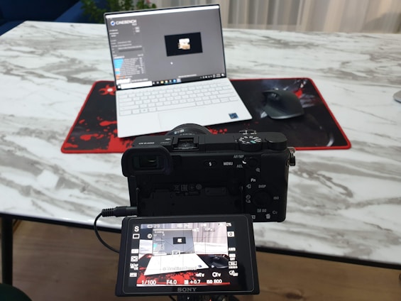 A camera positioned in front of a laptop on a marble-textured table. The camera's digital screen displays the laptop as well. A wireless mouse sits on a red and black mouse pad beside the laptop.