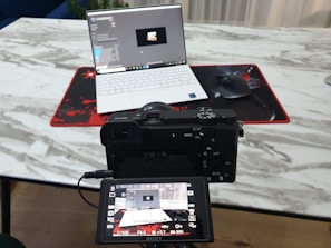A camera positioned in front of a laptop on a marble-textured table. The camera's digital screen displays the laptop as well. A wireless mouse sits on a red and black mouse pad beside the laptop.