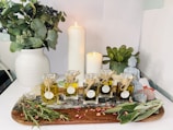 A serene arrangement featuring various essential oil bottles on a wooden tray surrounded by dried herbs and rose petals. A white and a slightly smaller candle are lit, casting a warm glow. A white vase filled with greenery stands on the left, and a small Buddha statue is placed near a potted succulent on the right. The setting suggests a tranquil atmosphere, possibly for meditation or relaxation.