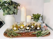 A serene arrangement featuring various essential oil bottles on a wooden tray surrounded by dried herbs and rose petals. A white and a slightly smaller candle are lit, casting a warm glow. A white vase filled with greenery stands on the left, and a small Buddha statue is placed near a potted succulent on the right. The setting suggests a tranquil atmosphere, possibly for meditation or relaxation.
