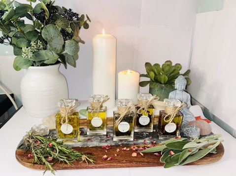 A serene arrangement featuring various essential oil bottles on a wooden tray surrounded by dried herbs and rose petals. A white and a slightly smaller candle are lit, casting a warm glow. A white vase filled with greenery stands on the left, and a small Buddha statue is placed near a potted succulent on the right. The setting suggests a tranquil atmosphere, possibly for meditation or relaxation.