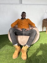 A person is sitting comfortably on a beige bean bag on a patch of green artificial grass. They are wearing an orange jacket, dark grey jeans, and tan work boots. The setting appears to be indoors with a minimalistic decor including a small wooden table to the side.