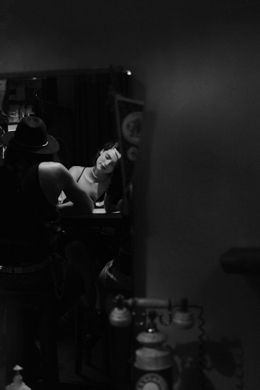 A monochromatic image captures two individuals seated at a table. One person is wearing a hat, and the other has their head tilted, resting their hand on the side of their face, appearing contemplative or tired. The lighting creates a moody and intimate atmosphere. There is an old-fashioned telephone visible in the lower part of the frame.