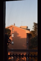 A window with vertical blinds partially drawn, allowing a view of a building bathed in orange sunlight. Shadows from trees are visible on the building's surface, and part of a clothes hanger or rack is silhouetted against the window.