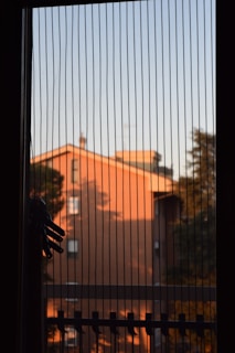 A window with vertical blinds partially drawn, allowing a view of a building bathed in orange sunlight. Shadows from trees are visible on the building's surface, and part of a clothes hanger or rack is silhouetted against the window.