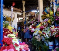 A bustling indoor flower market filled with vibrant and diverse blooms. People are seen interacting among the various colorful flower displays, with one person notably wearing traditional clothing and a bright woven shawl. The environment is lively with an abundance of flowers like roses, lilies, and sunflowers arranged in buckets and on tables.