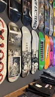 A variety of skateboard decks are displayed vertically on a wall. Each deck features unique designs and graphics, including black and white photography, colorful abstract patterns, and text. The decks are arranged in a row, showcasing their different artistic styles.